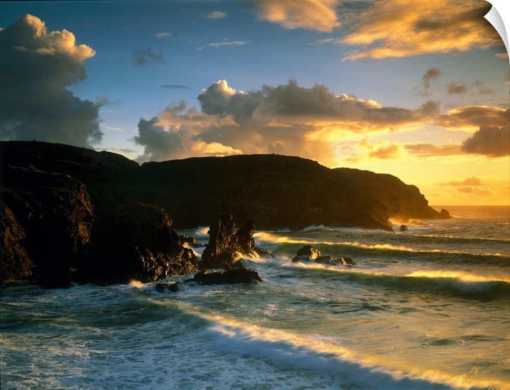 Photograph of rock cliffs in ocean with waves rolling in under a cloudy sky at sunrise.