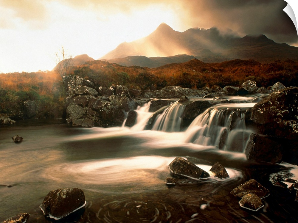 Time lapsed photograph of water flowing through a rocky river while the sun glows behind clouds in the distanced.