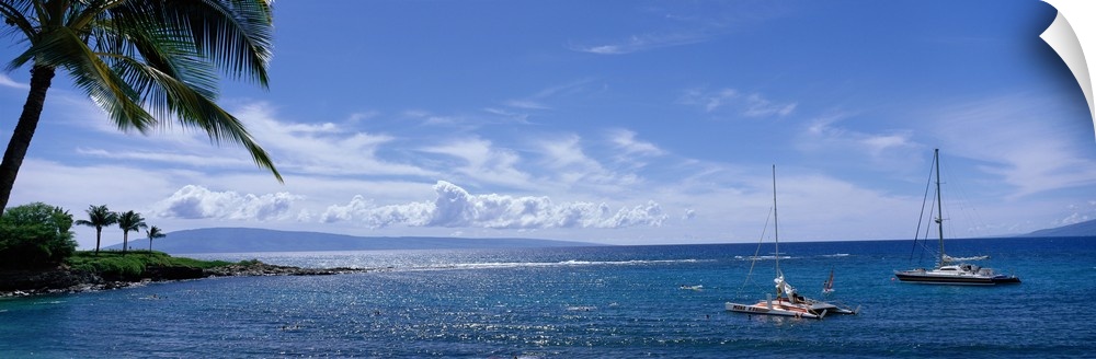 Panoramic photograph taken of a bay in Hawaii with land and palm trees on the left side and two boats anchored in the water.