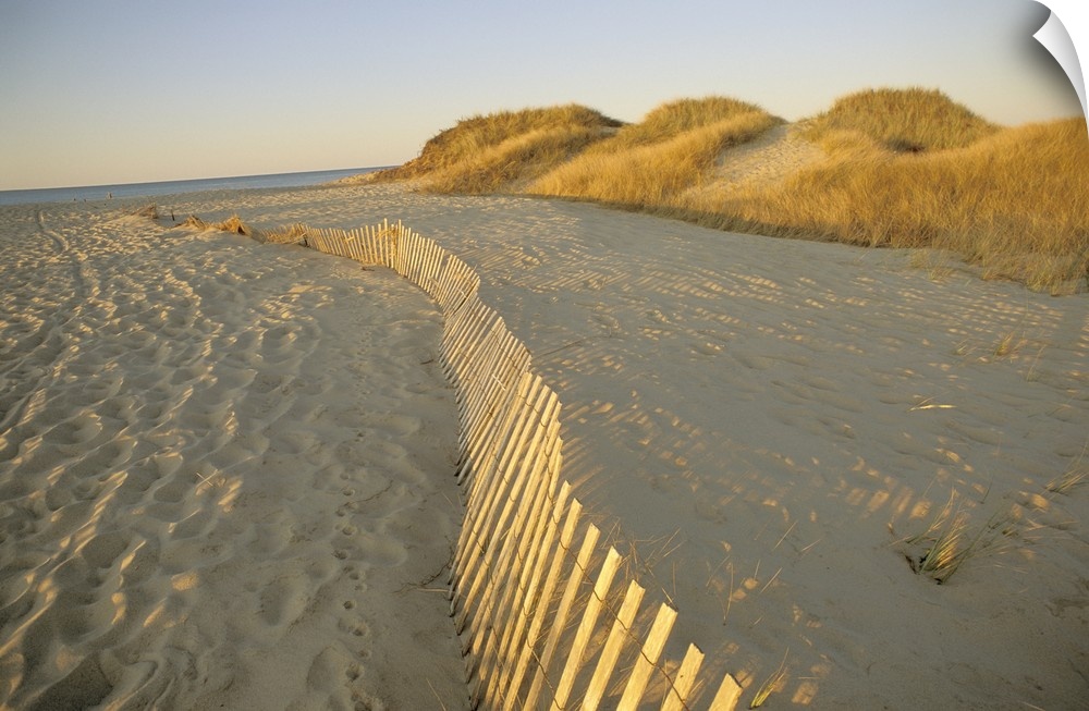 Large photograph taken of a sandy beach that has a fence running through the middle of the picture.  In the background the...