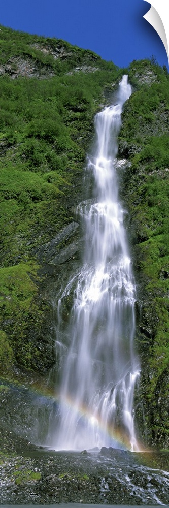 Vertical panoramic photograph of water cascading over rocks with small rainbow at the bottom.