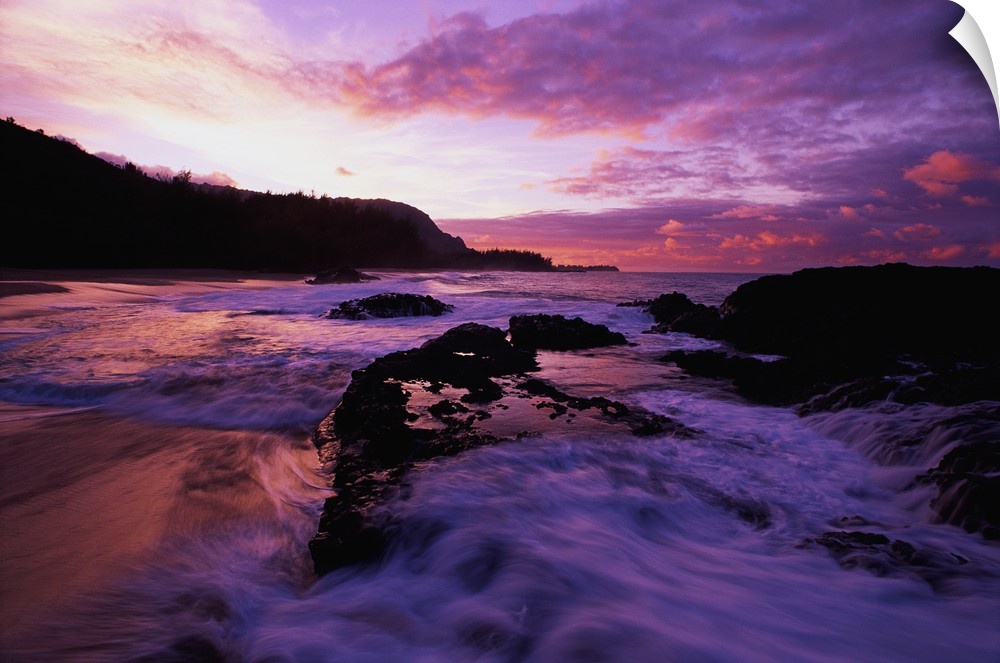 Photograph of rocky shoreline with waves rolling in at dusk.  There is a silhouette of a forest in the background.