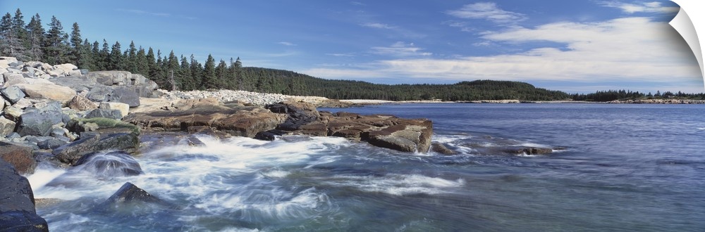 Panoramic photograph of rocky shoreline with surf.  There are trees in the distance under a cloudy sky.