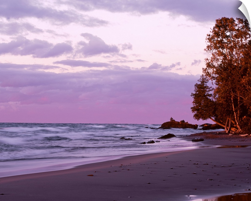 Picture of waves in Lake Superior crashing on to the beach as the sun sets and lights up the sky in purple tones.