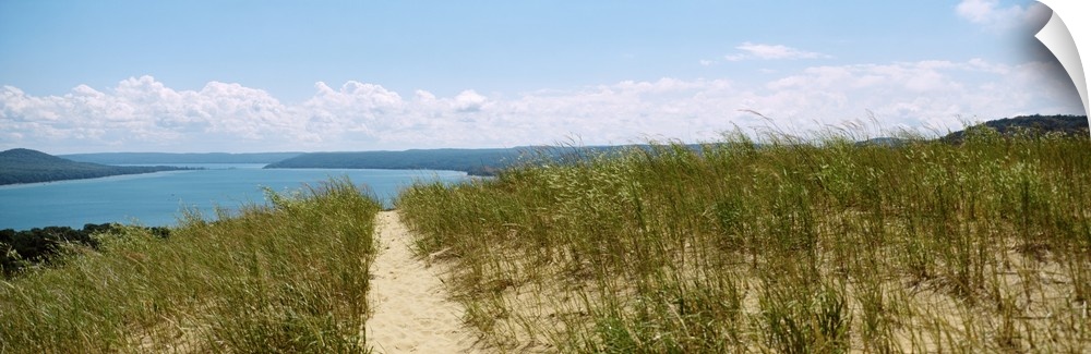 Panoramic photograph of grass covered beach with a sandy path leading to the ocean in the distance under a cloudy sky.