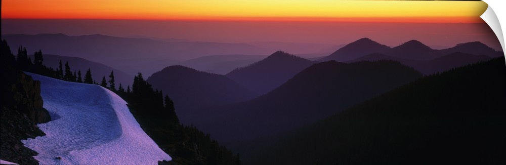 Mountains in Mount Rainier are silhouetted by the sunset in the background and captured in panoramic view.