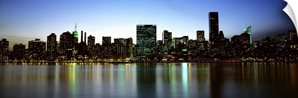 Panoramic photograph of the New York City skyline, lit at dusk and reflecting in the waters in the foreground.