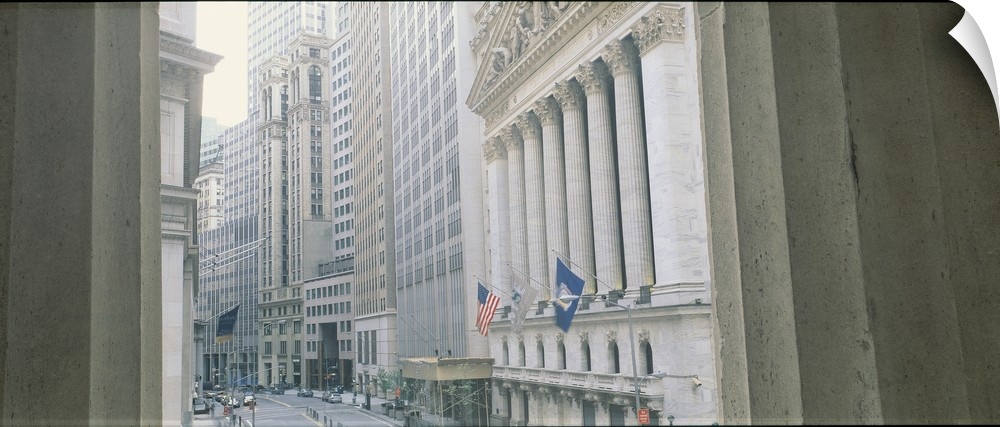 View between two concrete columns looking down Wall Street in New York City, with the faoade and columns of the Stock Exch...