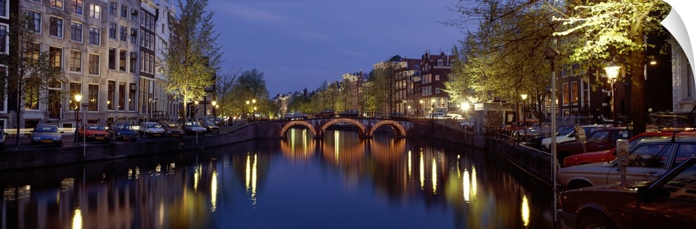 A wide angle view of a canal in Amsterdam with buildings and street lights illuminated on either side and reflecting in th...