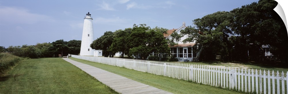 Horizontal photograph on a big canvas of a thin boardwalk and fence leading to a lighthouse surrounded by trees, a house i...
