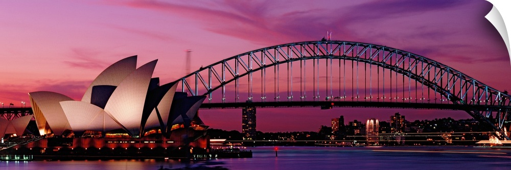 Panoramic photograph of iconic floating building structure with bridge and city skyline in distance lit up at sunset.