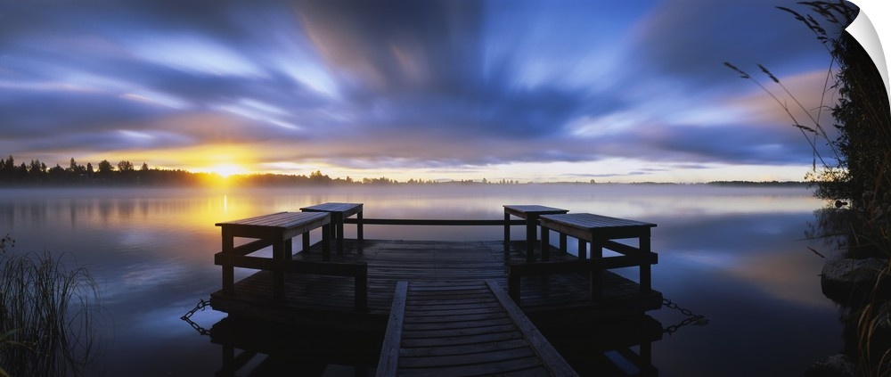 Panoramic photograph f wooden dock at sunset with forest in distance under a cloudy sky.