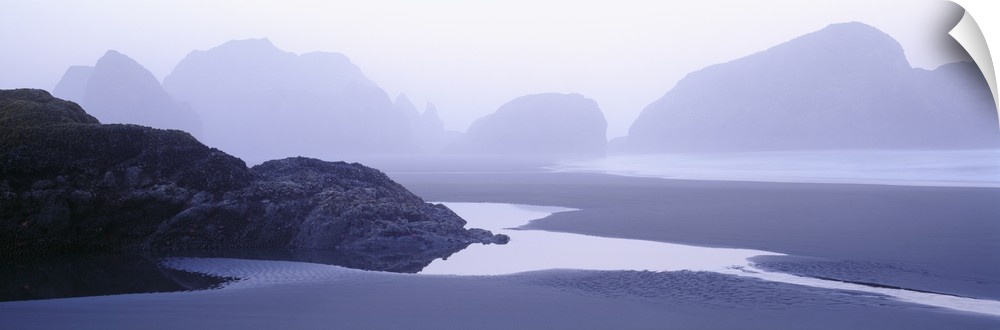 Rock formations in the water are faintly seen behind a layer of fog. Some water puddles on the beach near a smaller rock f...