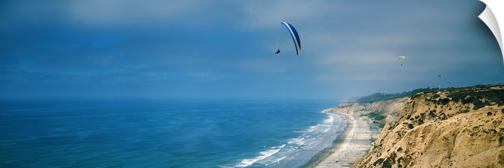 Horizontal photograph on a giant canvas of several paragliders floating over bright blue waters along the coast of La Joll...