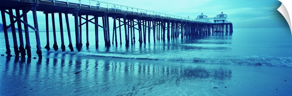 Monotone photograph of a long pier reaching out into the Pacific Ocean at dusk with shallow waves receding as the tide cha...