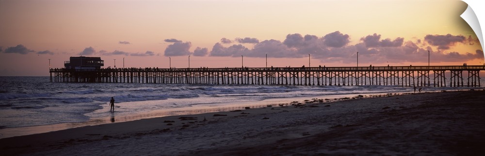 A beach board walk extends out into the ocean in this landscape photograph of the sun setting on a sandy beach.
