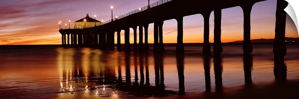 A panoramic photograph of the silhouette of a long pier over the ocean at sunset.