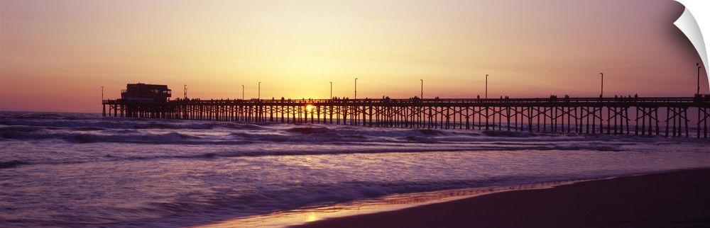 Panoramic photograph of the sunset that can barely be seen behind a long pier that reaches far out into the ocean.