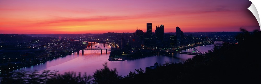 Oversized, landscape photograph of the distant skyline of Pittsburgh, Pennsylvania lit up, beneath a vibrant sunset.