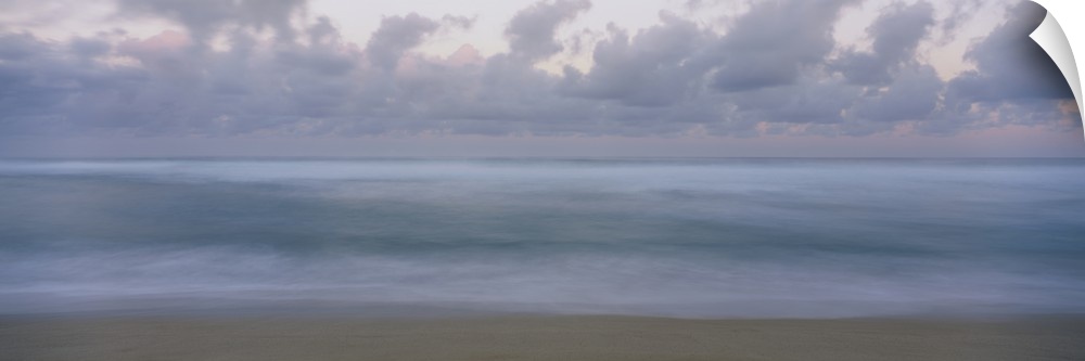 This is a panoramic photograph of a waves gently washing on the sandy shore with a cloudy sky overhead in this seascape.