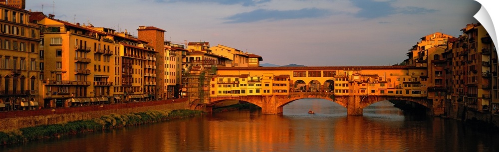 Wide angle photograph of buildings alongside the Arno River, with a view of he Ponte Vecchio bridge, at sunset in Florence...
