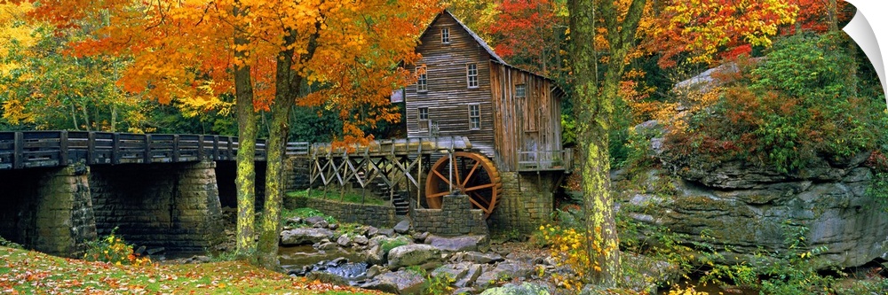 Panoramic picture taken of a mill through autumn colored trees with a bridge just to the left.