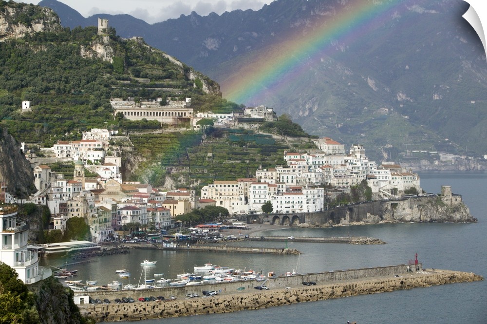 This is a picture of a rainbow over a town off a coast in Italy. Large white buildings line the water that has piers with ...