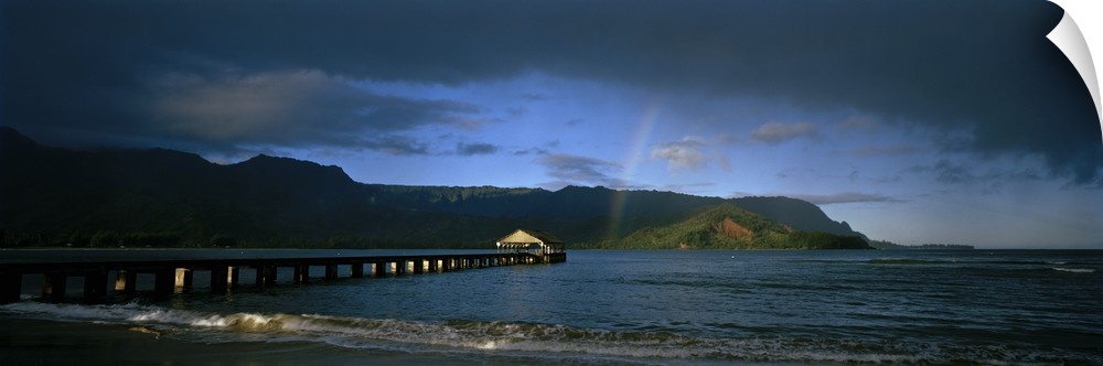 Picture taken of a long pier that reaches far out into the ocean with a faint rainbow shown near land that is in the dista...