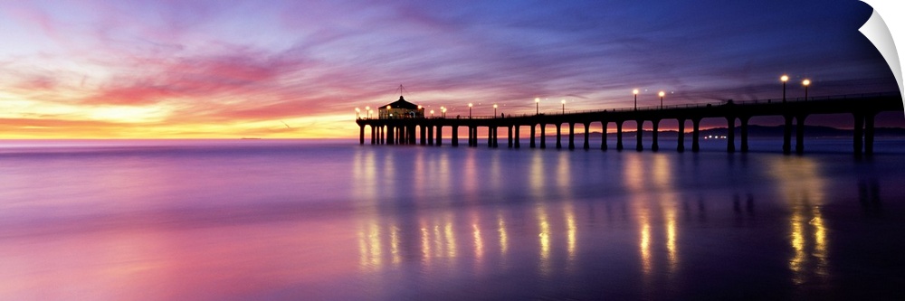 Panoramic photograph of pier extending into ocean at sunset. The lights on the pier are reflected in the ocean and the sky...