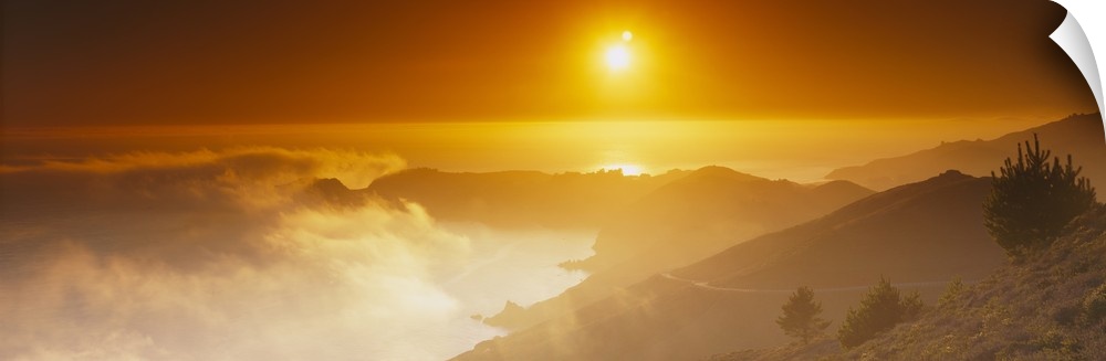 Panoramic photo on canvas of the sun shining bright in the sky above an ocean and mountains with low clouds on the left.