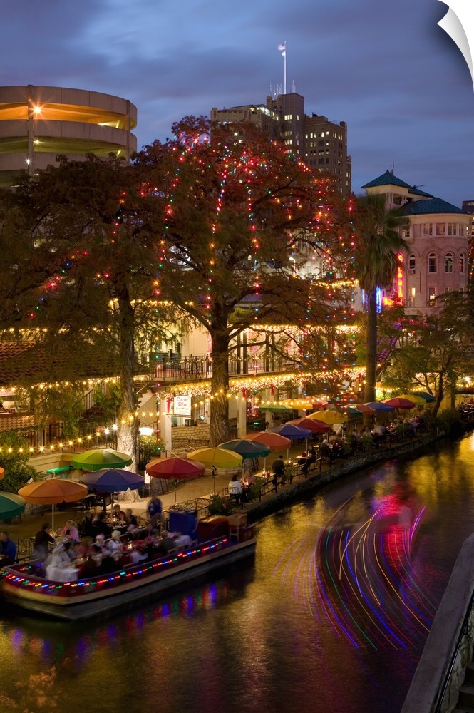 Vertical photo on canvas of people eating at an outdoor restaurant next to a river underneath umbrellas and sparkling lights.