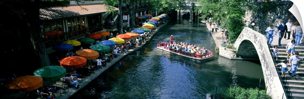 A panoramic picture of a river in Texas that is lined with tables that have colorful umbrellas and a small passenger boat ...