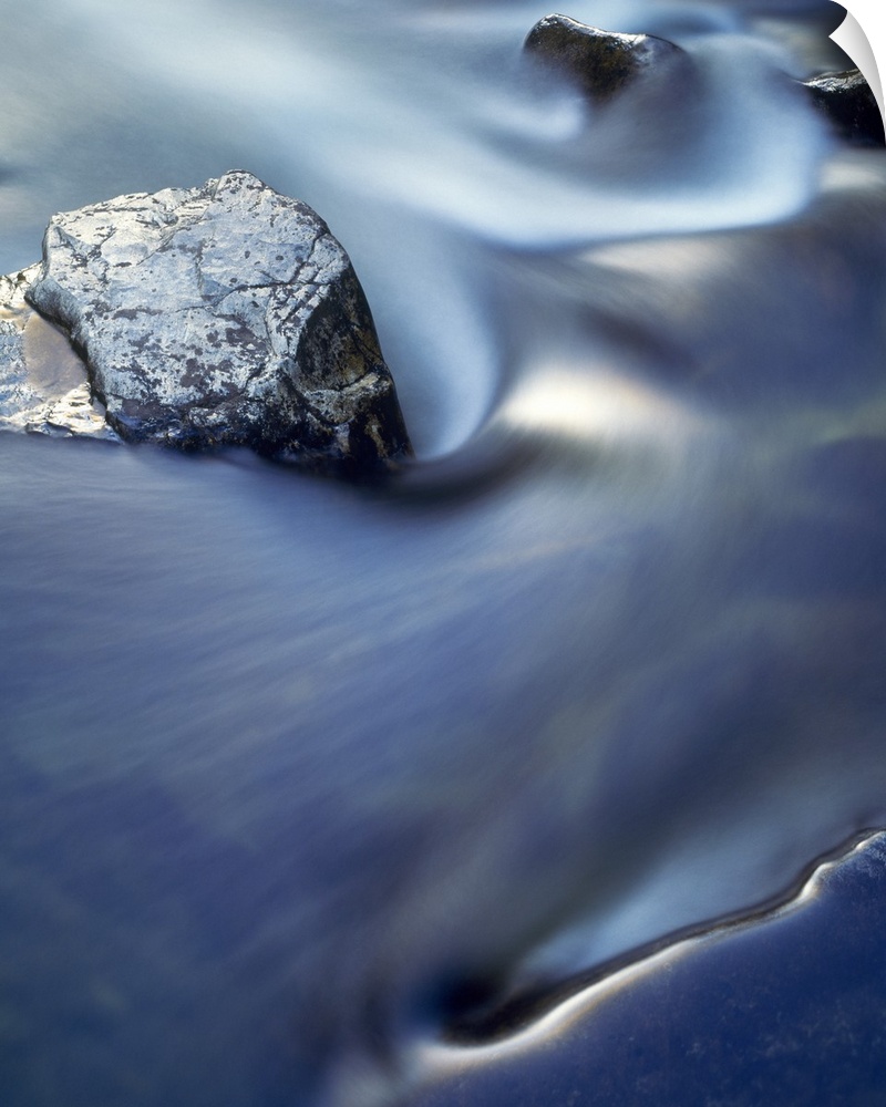 Vertical, close up photograph on a large wall hanging  of blurred water in rapid movement as it swirls around rocks in the...