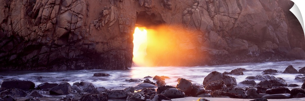 Panoramic photograph of rocky shoreline with sun peering through huge stone arch-like structure.