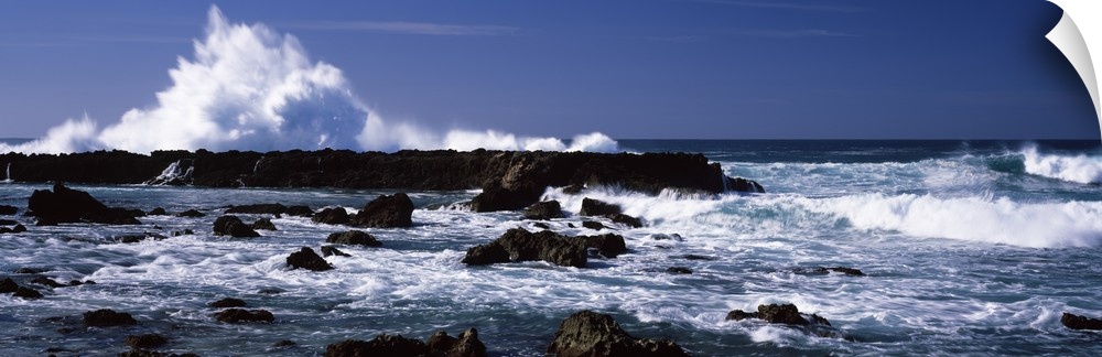 Panoramic photograph of rocks at sea with waves crashing around them under a clear sky.