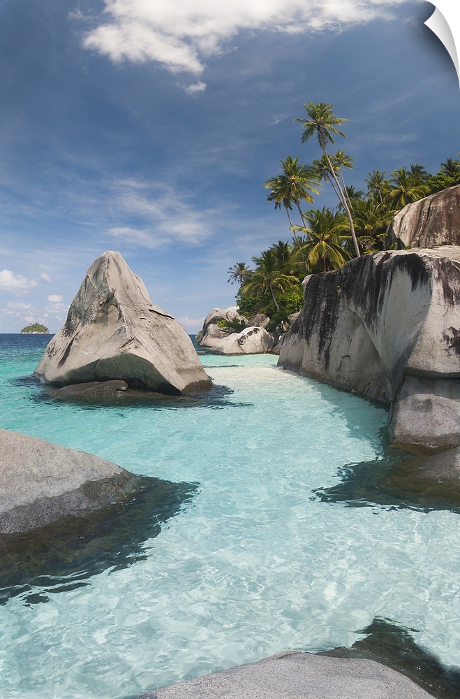 Shallow waters at the edge of a tropical beach full of large stone boulders and palm trees.