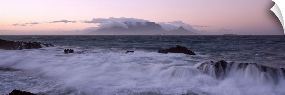 Waves from the rising tide crash over the rocky coastline at twilight. In the distance clouds form over mountain peaks.