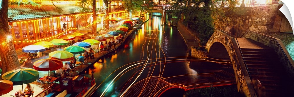 Nighttime panoramic photo of the Riverwalk in San Antonio as boats travel through the canals and people eat under colorful...