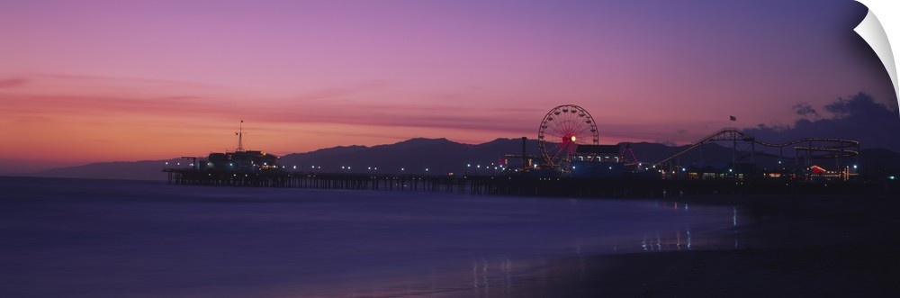 A panoramic photograph of a board walk, Ferris wheel, and rollercoaster on a board walk during a pastel sunset.
