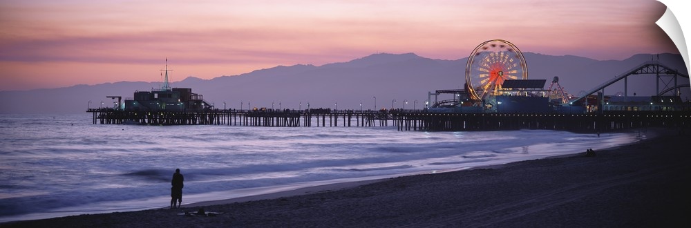 A panoramic shot taken of the Santa Monica pier during sun down with a couple standing on the beach at the waters edge.