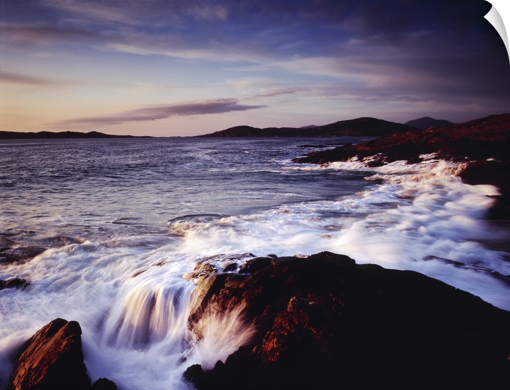 Horizontal photograph of water crashing into the rocky shoreline of Outer Hebrides as the sun sets over Scotland, a mounta...