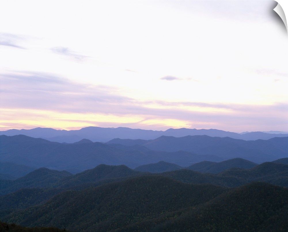 Rows of silhouetted mountains covered in green leaf trees as far as the eye can see as the sun rises in the distance.