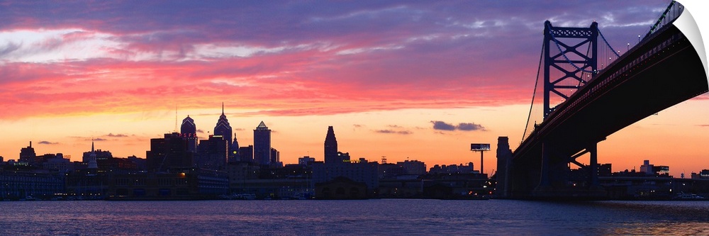 View from the shoreline under the bridge of pastel-colored clouds over the city skyline at dusk.