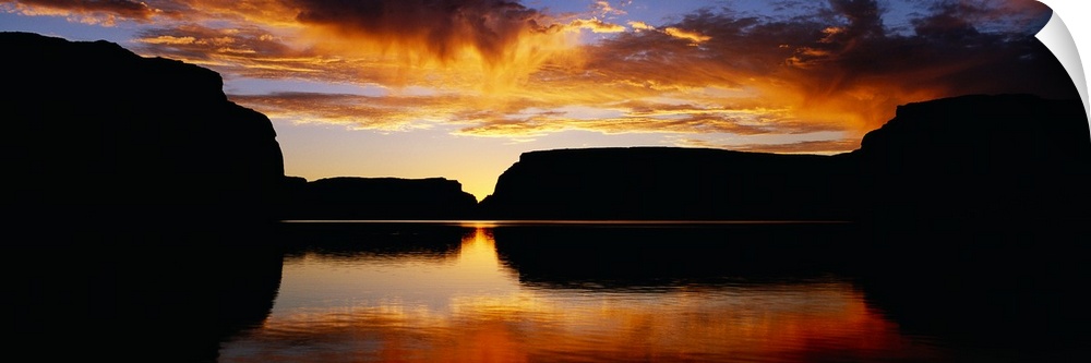 Giant landscape photograph of large, silhouetted rocks surrounding Lake Powell, beneath a vibrant sky at sunset, in Utah.