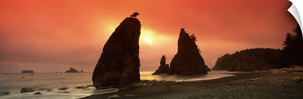 This is a panoramic photograph of the sun setting behind enormous rock formations on this rocky Northwestern beach.