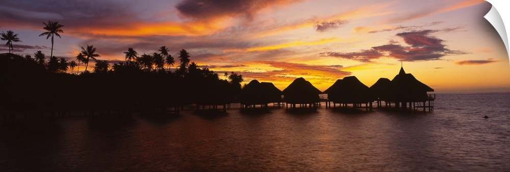 Long and narrow photo on canvas of palm trees and houses on stilts over the ocean silhouetted against a bright sunset.