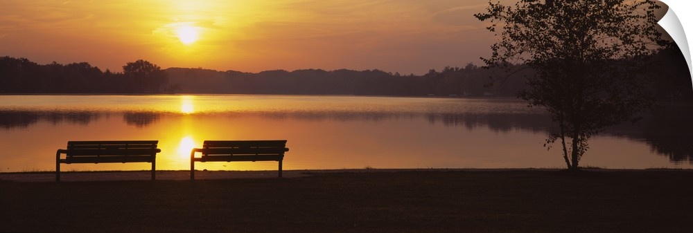Panoramic photograph of park benches near water's edge at sunset with forest silhouette in the distance.