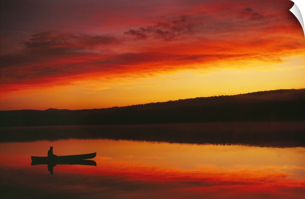 Big, landscape photograph of the silhouette of a person sitting in a boat beneath a vibrant, fiery sunset.