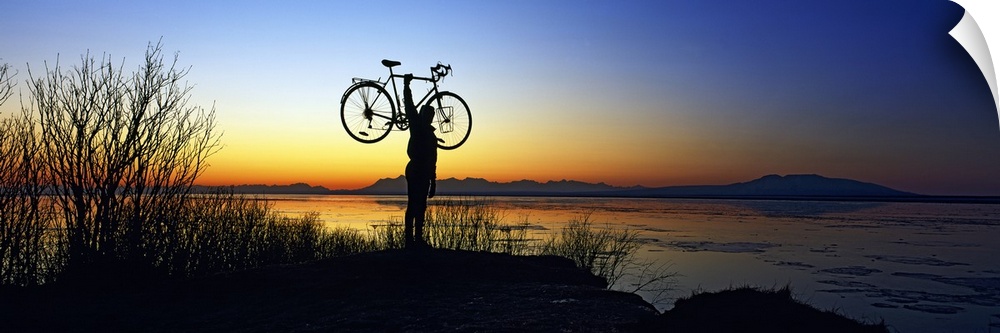 Panoramic photograph of man holding bike in air at shoreline at dusk with mountains in the distance.