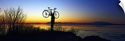 Silhouetted cyclist holding bicycle over head, river's edge, sunset, Alaska